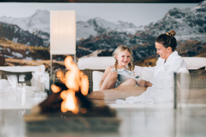 eine Mama und ihre Tochter sitzen gemeinsam im Bademantel im Wellnessbereich, im Vordergrund brennt ein beruhigendes Feuer und im Hintergrund sieht man die verschneite Bergkulisse