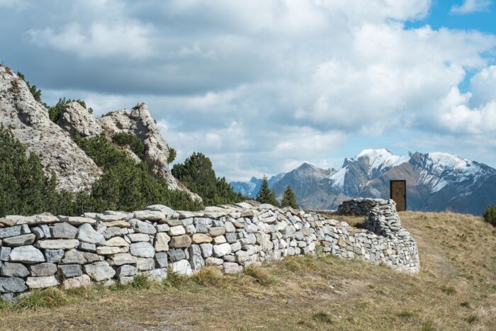 Steinmauer mit einer Türe am Ende der Mauer; im Hintergrund erstreckt sich das Bergpanorama