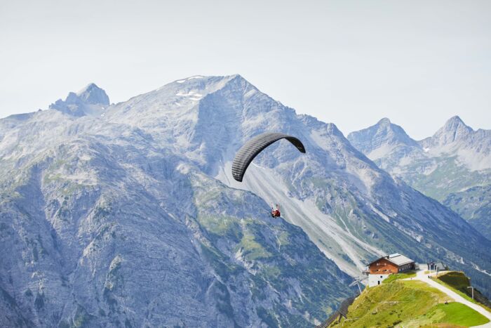 Ein Paragleiter von hinten zu sehen und im Hintergrund eine große weite zu sehen mit Berggipfeln
