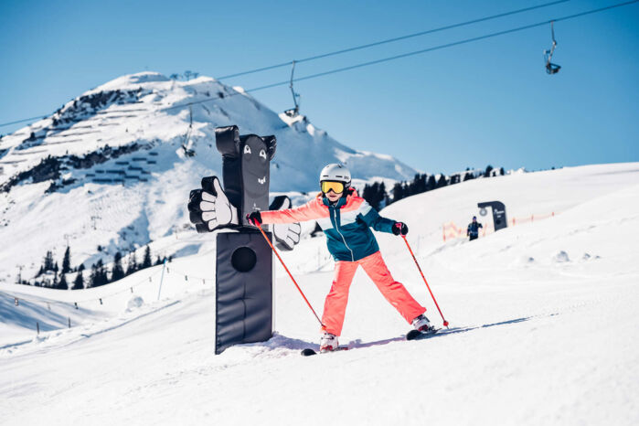 Junge Skifahrerin schlägt lachend gegen eine Daumen-hoch-Figur an einer Spielstation im Skigebiet Lech, umgeben von Bergpanorama und Sessellift.