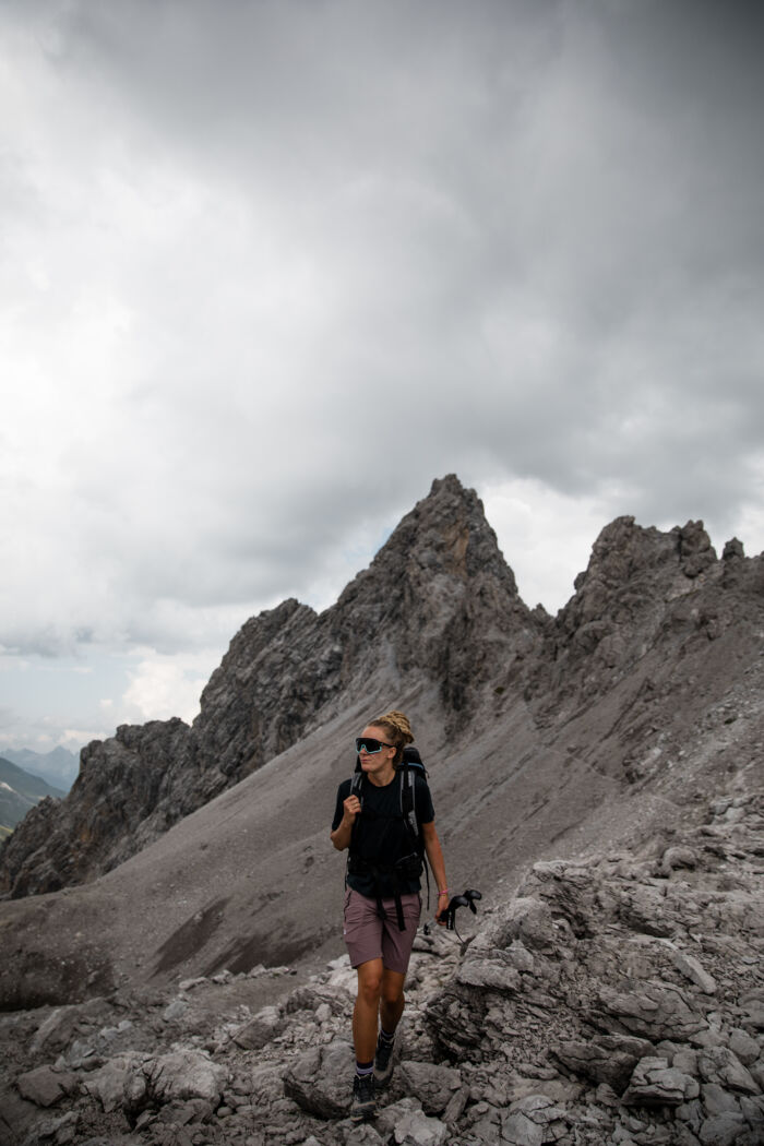 Wanderin mit Rucksack steht auf einem felsigen Grat vor schroffen Gipfeln unter bewölktem Himmel.