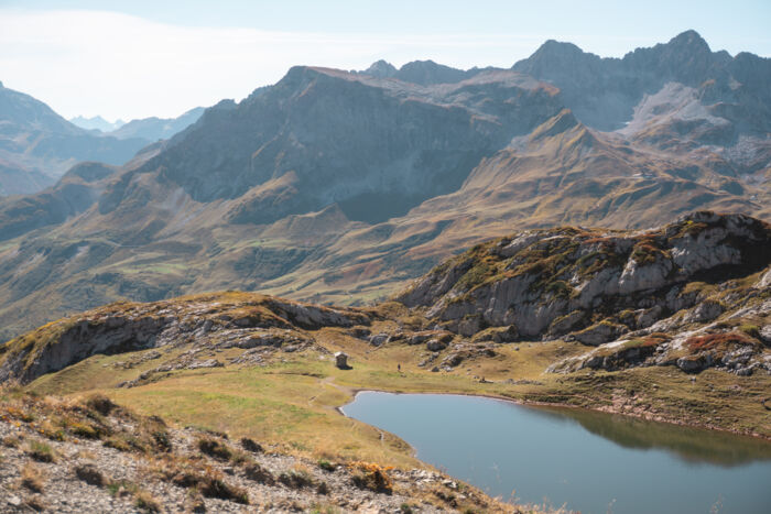 Blick auf einen kleinen Bergsee mit einer Steinhütte am Ufer, umgeben von herbstlich gefärbten Almwiesen und Felsen, im Hintergrund mehrere dunstige Bergketten.
