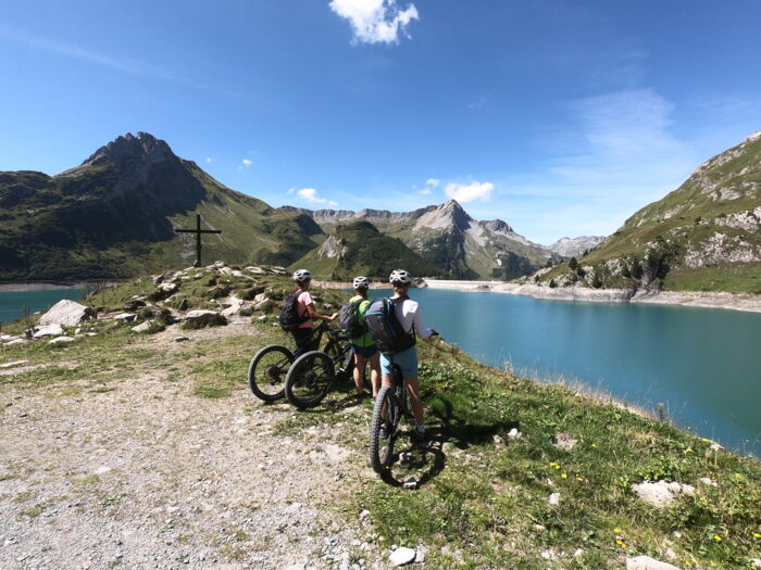 Drei Radfahrer stehen an einem Aussichtspunkt und blicken vorbei an einem Gipfelkreuz über einen türkiesen See auf ein wunderschönes Bergpanorama.