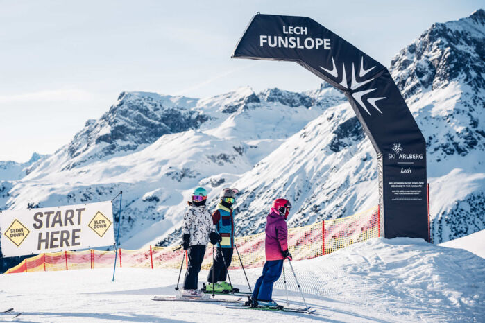 Drei Skifahrer stehen am Startbogen der Lech Funslope, umgeben von schneebedeckten Bergen.
