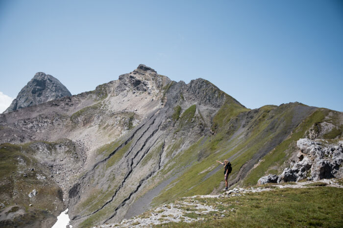 Wanderin zeigt auf einen fernen Gipfel, während sie auf einem grünen Grat mit Blick auf schroffe Berge steht.
