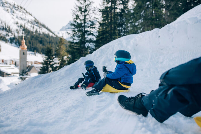 zwei Kinder in Skigewand rodeln mit dem Zipfelbob die Rodelbahn in Lech Zürs hinunter, im Hintergrund sieht man die Kirche von Lech
