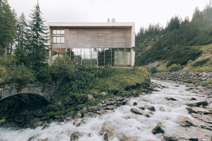 Biomasseheizkraftwerk Zürs ist ein Holzgebäude von der Hinteransicht aus Holz und Glasfassade und nebenan sieht man den Bach mit Wasser