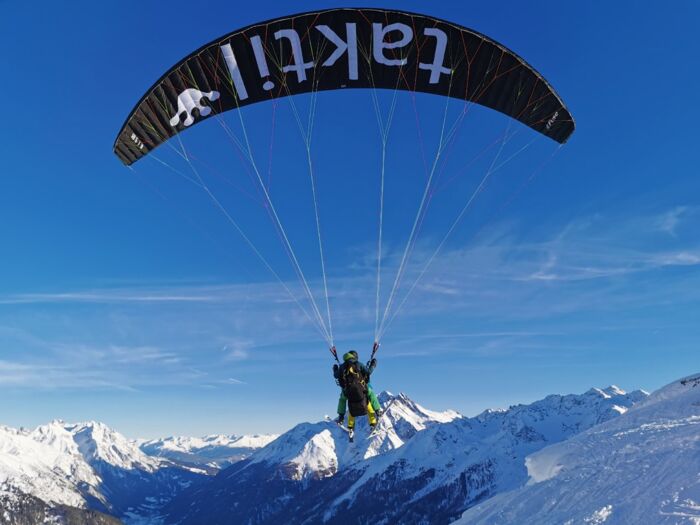 Gleitschirm mit Tandemflug zu sehen von hinten hängen zwei Personen am Gleitschirm und im Hintergrund ist die Berglandschaft zu sehen