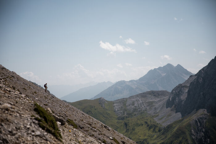 Einzelner Wanderer steht klein auf einem steinigen Hang, im Hintergrund weite Bergketten im Dunst.