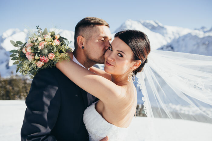 Bräutigam küsst seine Braut auf die Wange, im Hintergrund ragen die verschneiten Berge hoch