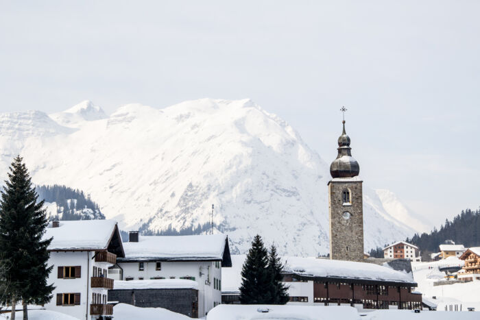 Verschneite Landschaft im Hintergrund und im Vordergrund die Kirche sowie zwei Häuser in Lech zu sehen