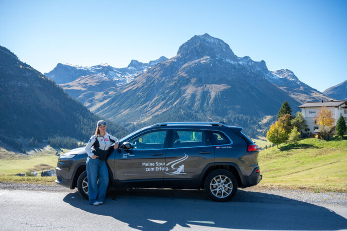 Skirennläuferin Leonie Zegg posiert vor ihrem Auto und im Hintergrund ist eine Berglandschaft zu sehen. Unter anderem ist auf dem Auto ein Aufdruck zu sehen mit: Meine Spur zum Erfolg