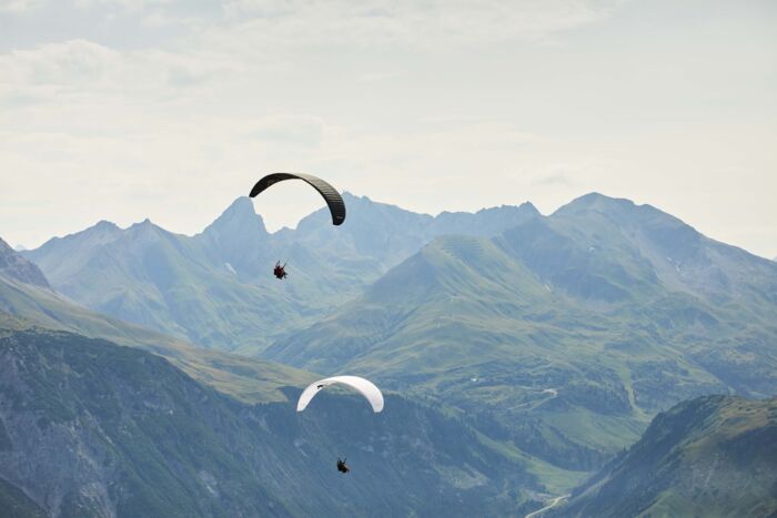 Zwei Paragleiter von hinten zu sehen und im Hintergrund eine große weite zu sehen mit Berggipfeln