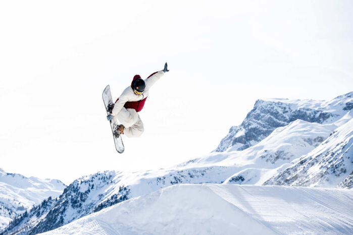Freestyler springt über eine Schanze und im Hintergrund ist strahlend blauer Himmel und eine wunderschöne Bergkulisse zu sehen.