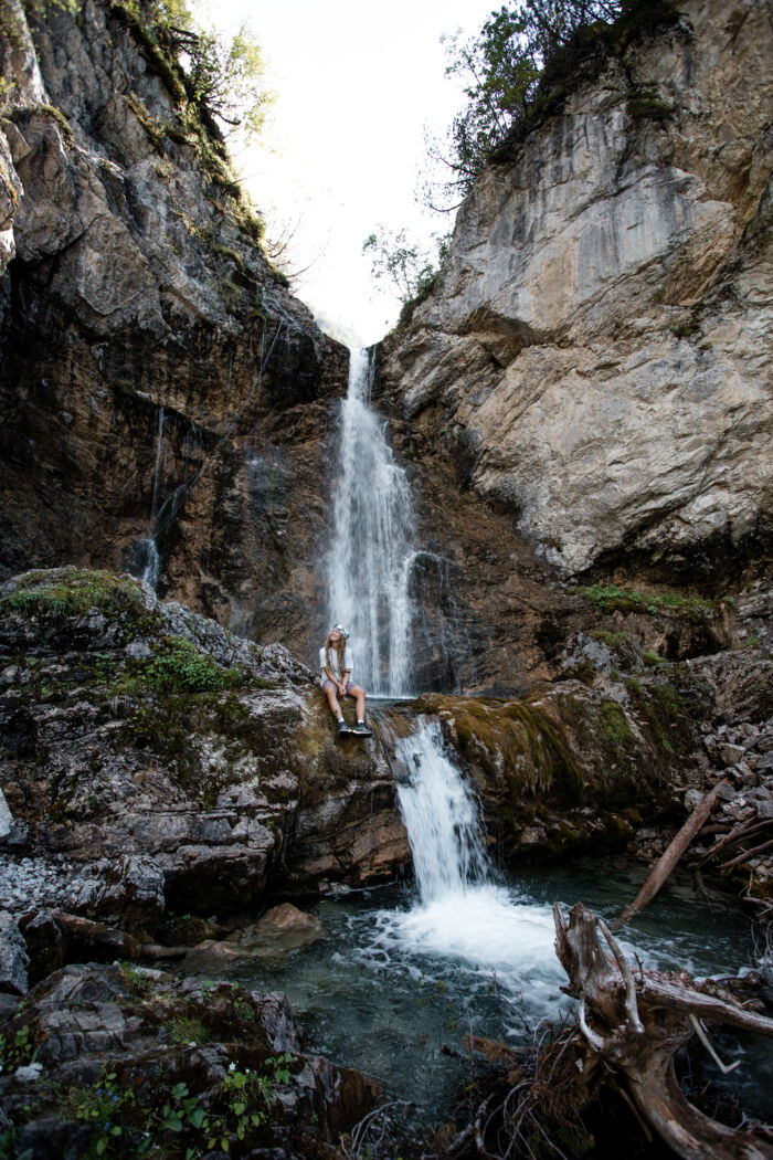 Wanderin sitzt auf einem Felsvorsprung vor einem hohen Wasserfall in einer engen Schlucht.