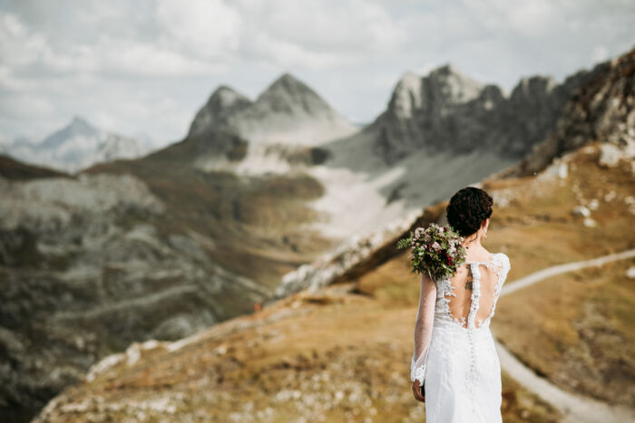 Braut steht von hinten sehend vor herrlicher Berglandschaft mit Blumenstrauß in der Hand