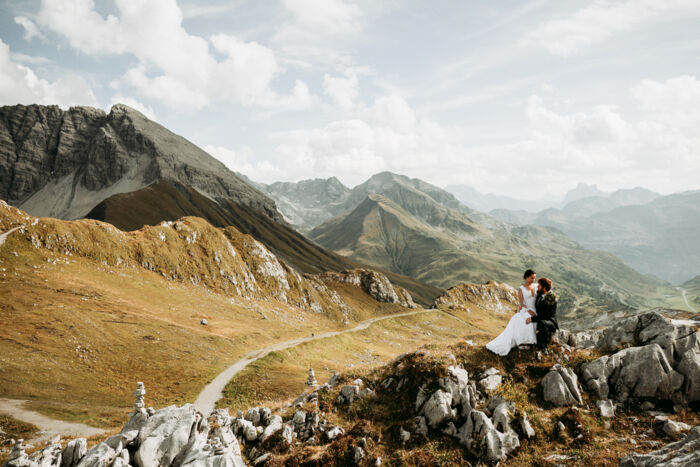 Frisch vermähltes Ehepaar sitzt auf Hügel vor traumhafter Berglandschaft und schaut sich in die Augen