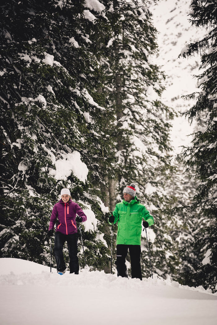 Blick auf die verschneiten Wälder, während im Vordergrund zwei Personen beim Schneeschuhwandern sind