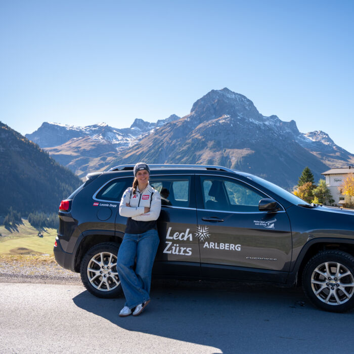 Frau steht vor Auto im Hintergrund ist das Omeshorn, der Hausberg von Lech zu sehen. Auf dem Auto ist ein großer Sticker-Aufdruck mit Lech Zürs Stern Arlberg zu sehen