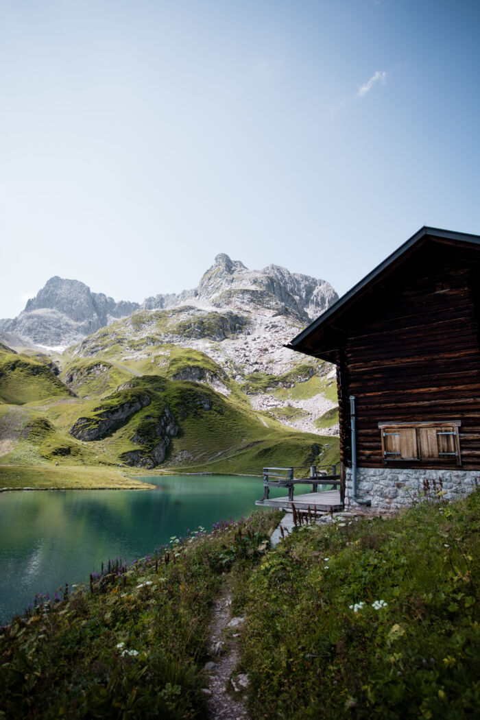 Almhütte am Ufer eines türkisfarbenen Bergsees, im Hintergrund felsige Gipfel.