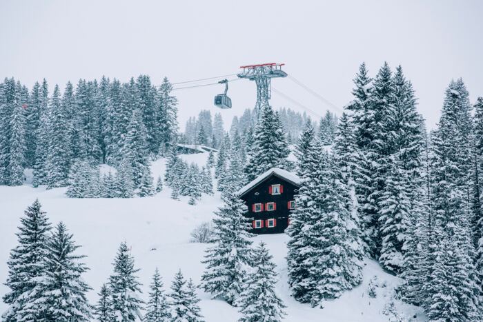 die Oberlecher Bergbahn fährt über die traumhafte Schneelandschaft, im Vordergrund ragt ein uriges Holzhaus zwischen den verschneiten Tannenbäumen heraus