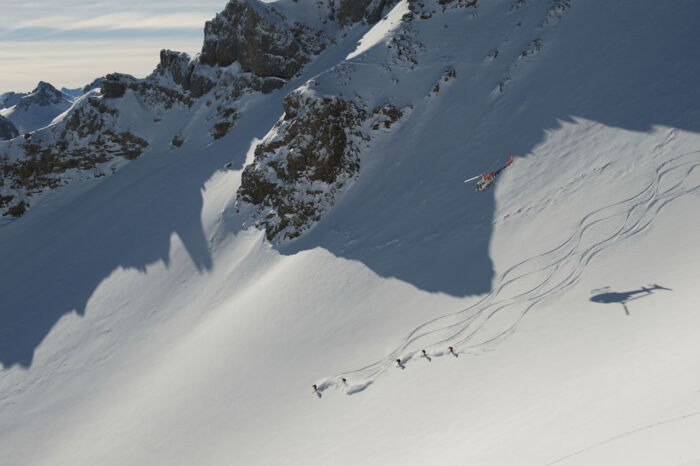 Blick auf eine Gruppe beim Heliskiing, die die ersten Schwünge in der unbefahrenen Offpiste fahren, über ihnen fliegt der Helikopter