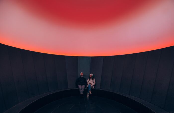 zwei Personen betrachten das Lichtspektakel im Skyspace, durch die Sonneneinstrahlung erscheint es in einem rötlichen Farbspiel