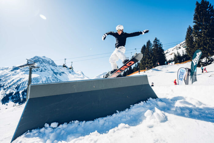 Freestyler fährt über eine Rail und im Hintergrund ist strahlend blauer Himmel und eine wunderschöne Bergkulisse zu sehen.