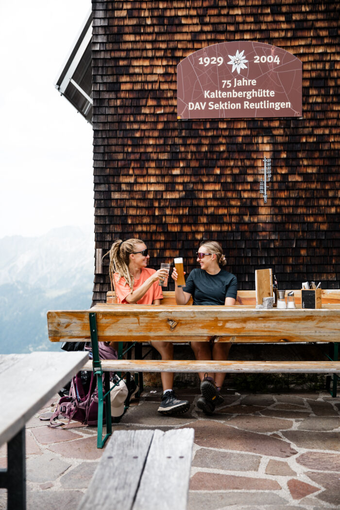 Zwei Wanderinnen stoßen mit Bier an einem Tisch vor der Kaltenberghütte an, im Hintergrund Berge.