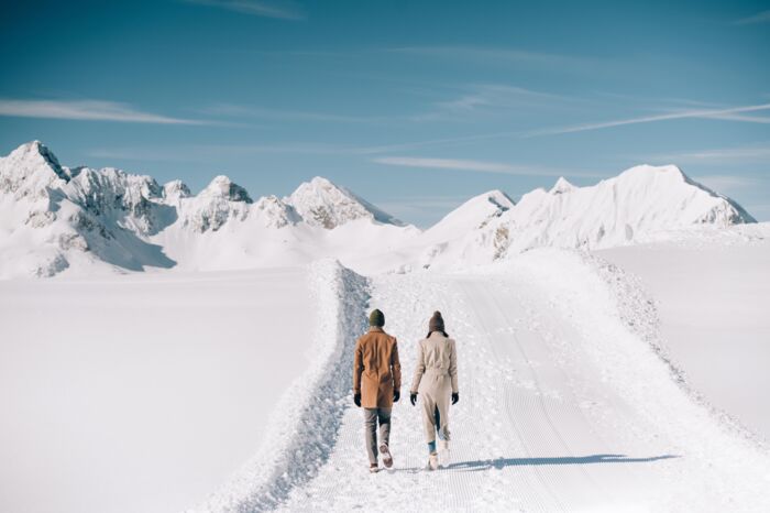 Ein Pärchen macht einen romantischen Spaziergang am Schneeflockenpanoramaweg bei klarem blauem Himmel und mit den verschneiten Berggipfeln im Hintergrund