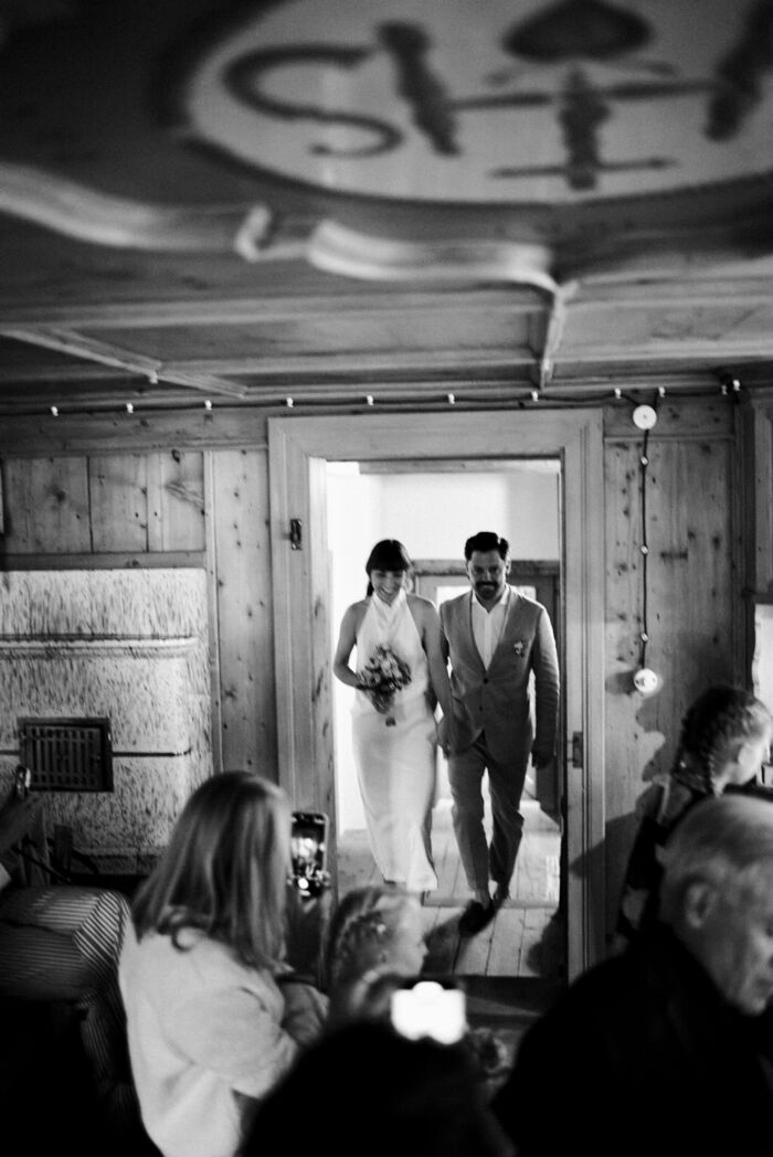 Hochzeitspaar läuft durch die Türe im Innenraum vom Huberhus auf dem Weg an der sitzenden Hochzeitsgesellschaft vorbei zur Trauung