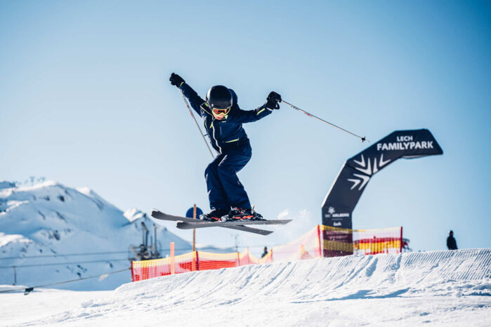 Junger Skifahrer springt mit gekreuzten Ski über einen Kicker im Lech Familypark, im Hintergrund Berge und blauer Himmel.