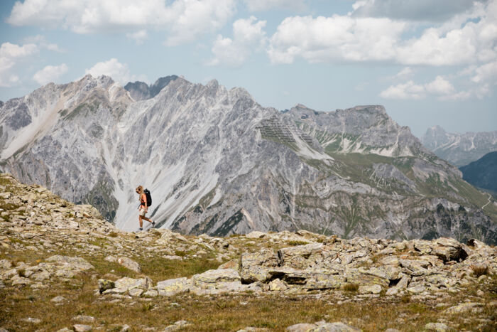 Wanderin mit Rucksack geht über einen felsigen Bergrücken, im Hintergrund schroffe Gipfel der Lechtaler Alpen.
