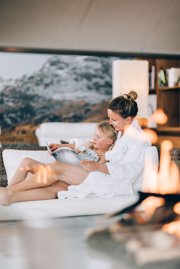eine Mama liest ihrem Kind eine Geschichte im Wellnessbereich vor im Hintergrund sieht man die atemberaubenden Berggupfel