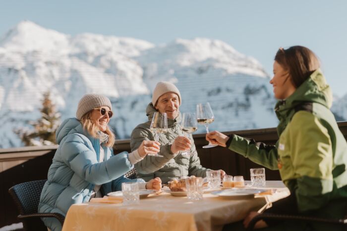 drei Freunde genießen auf der Sonnenterrasse die winterliche Berglandschaft und die Kulinarik von Lech Zürs mit einem Glas Wein