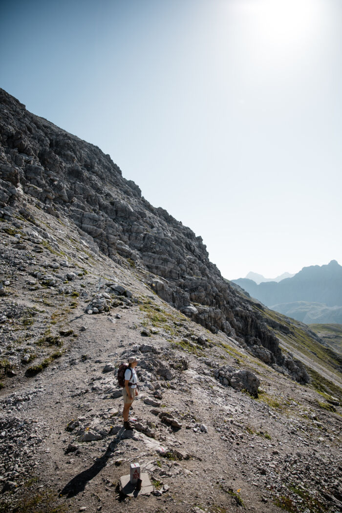 Wanderer steht an einem Wegpunkt mit Steinmarkierung am kargen Berghang, im Hintergrund weitere Gipfel.