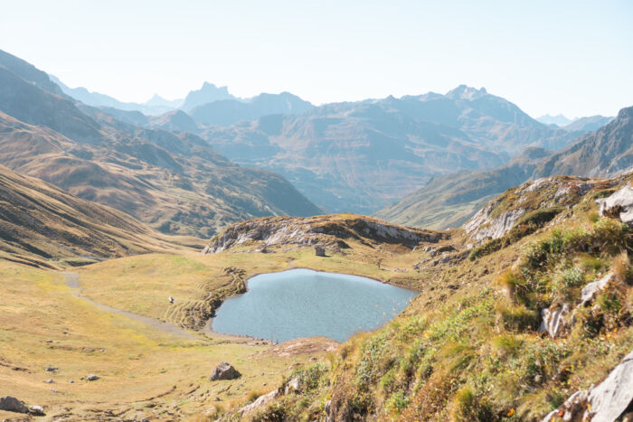Blick auf einen kleinen Bergsee mit einer Steinhütte am Ufer, umgeben von herbstlich gefärbten Almwiesen und Felsen, im Hintergrund mehrere dunstige Bergketten.