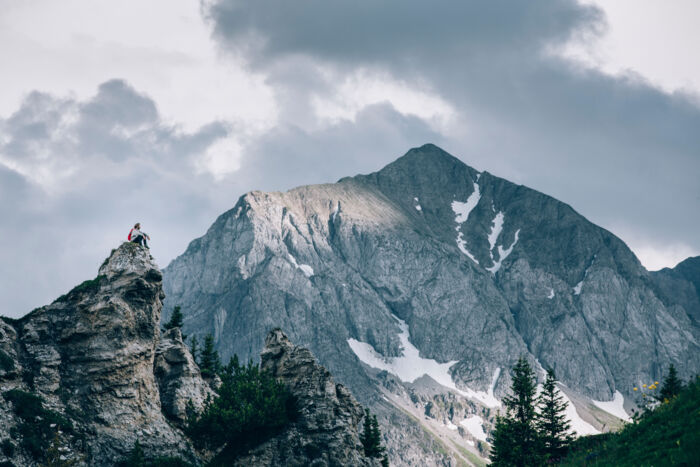ein Wanderer sitzt auf einem Felsvorsprung in Oberlech und genießt die Aussicht, im Hintergrund ist ein felsiger Berg mit Schneefeldern zu sehen