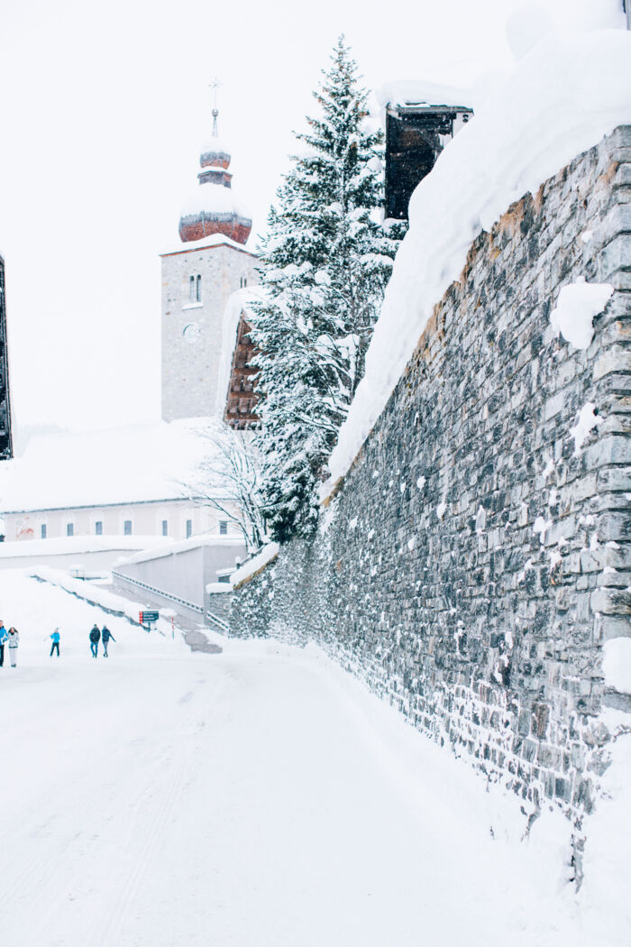 Blick vom schneebedeckten Gehweg auf die Kirche in der Dorfmitte von Lech, davor spazieren einige Personen durch den Schnee