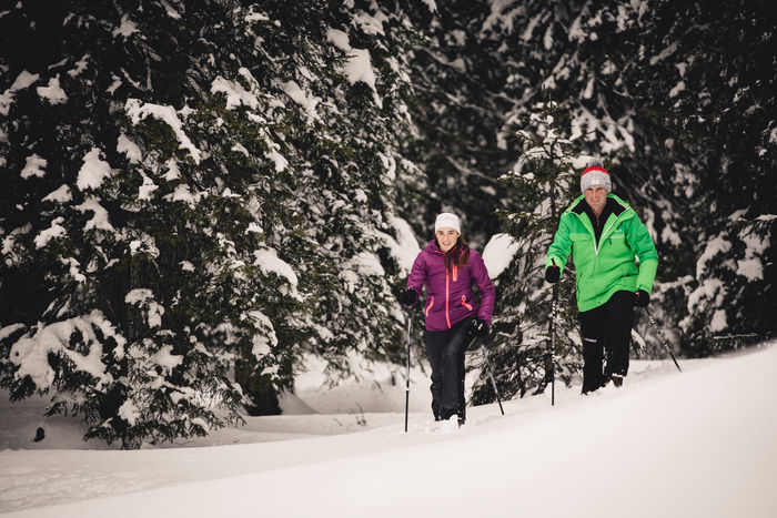 zwei Personen wandern durch die Schneelandschaft, im Hintergrund sieht man die verschneiten Nadelbäume