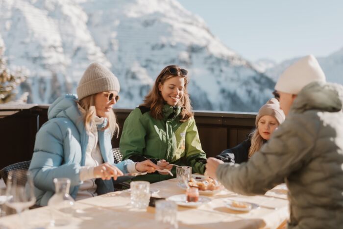 eine Familie genießt den warmen Kaiserschmarren auf der Sonnenterrasse mit einem atemberaubenden, verschneiten Bergpanorama in Lech Zürs am Arlberg