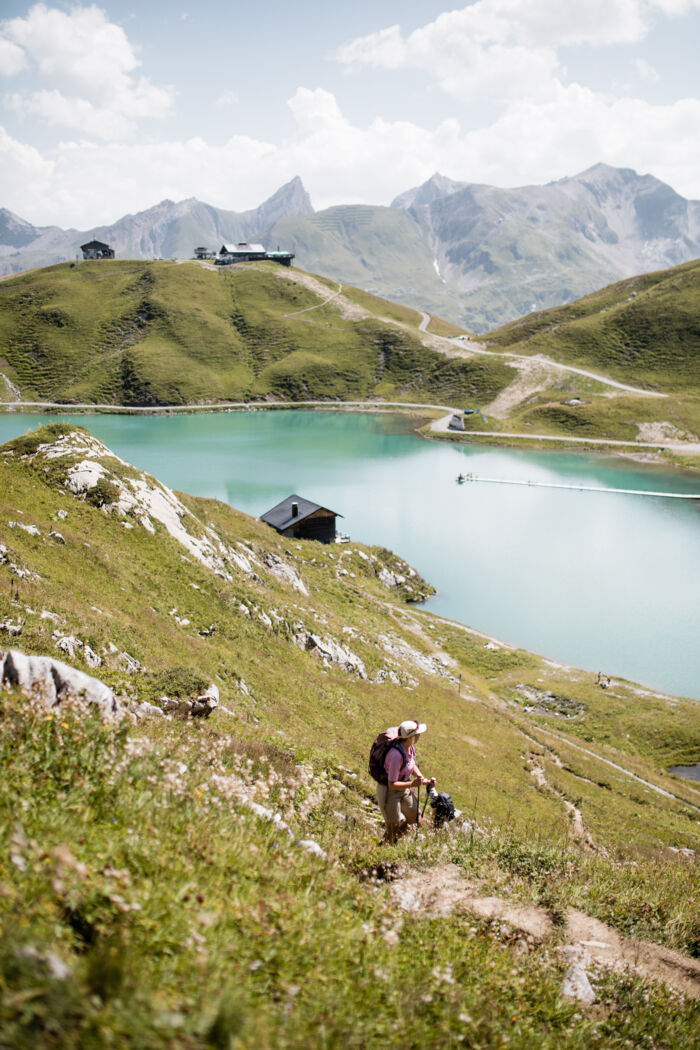 Wanderin mit Trekkingstöcken steigt einen grünen Hang hinauf, im Hintergrund der türkisfarbene Zürsersee.