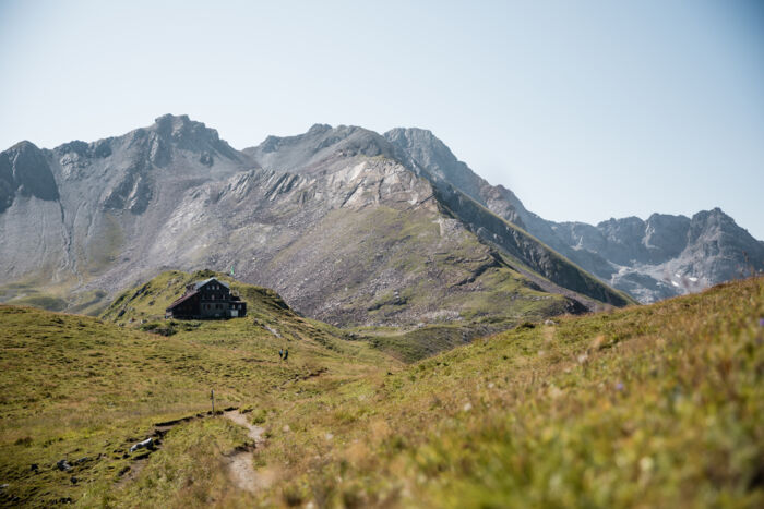 Die Stuttgarter Hütte liegt auf einer grünen Bergkuppe vor schroffem Felsmassiv, ein Wanderweg führt darauf zu.