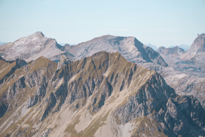 Zerklüftete Bergkette im Herbst mit felsigen Graten und herbstlich gelbbraunen Grashängen, dahinter hellere, graue Gipfel unter einem klaren, hellblauen Himmel.