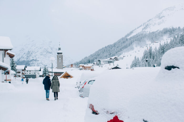 Blick auf das schneebedeckte Lech während zwei Personen über den eingeschneiten Gehweg spazieren und im Hintergrund die winterliche Bergkulisse erscheint