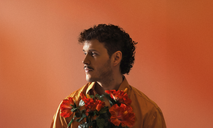 Der Künstler Marten mit lockigem Haar und Schnurrbart blickt im Profil zur Seite und hält rote Hibiskusblüten vor einer orangefarbenen Wand.