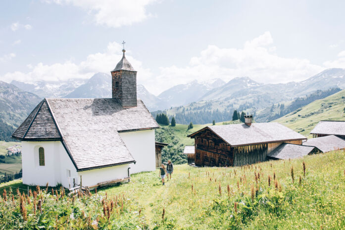 Blick auf die Kirche und urige Holzhäuser in Bürstegg, während zwei Wanderer auf dem Wanderweg laufen, im Hintergrund ist das leicht bewölkte Bergpanorama zu sehen