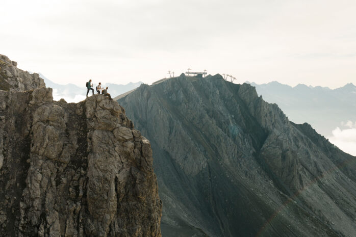 Wanderer sitzen und stehen an einem abschüssigen Felsvorsprung; im Hintergrund befindet sich ein Gebirgshang mit der Bergstation einer Seilbahn