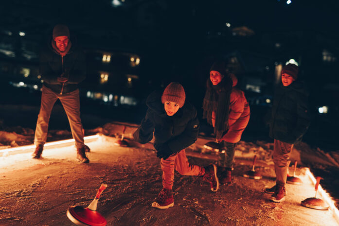 Die gesamte Familie ist auf dem Eisstockbahn und der jüngste Sohn wirft gerade den Eisstock nach vorne auf die Bahn alle schauen gespannt