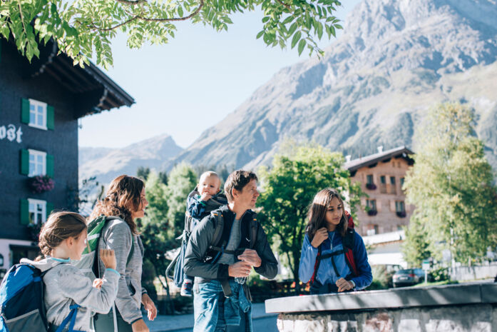 eine Familie steht vor dem Dorfbrunnen in Lech, im Hintergrund sieht man das Omeshorn